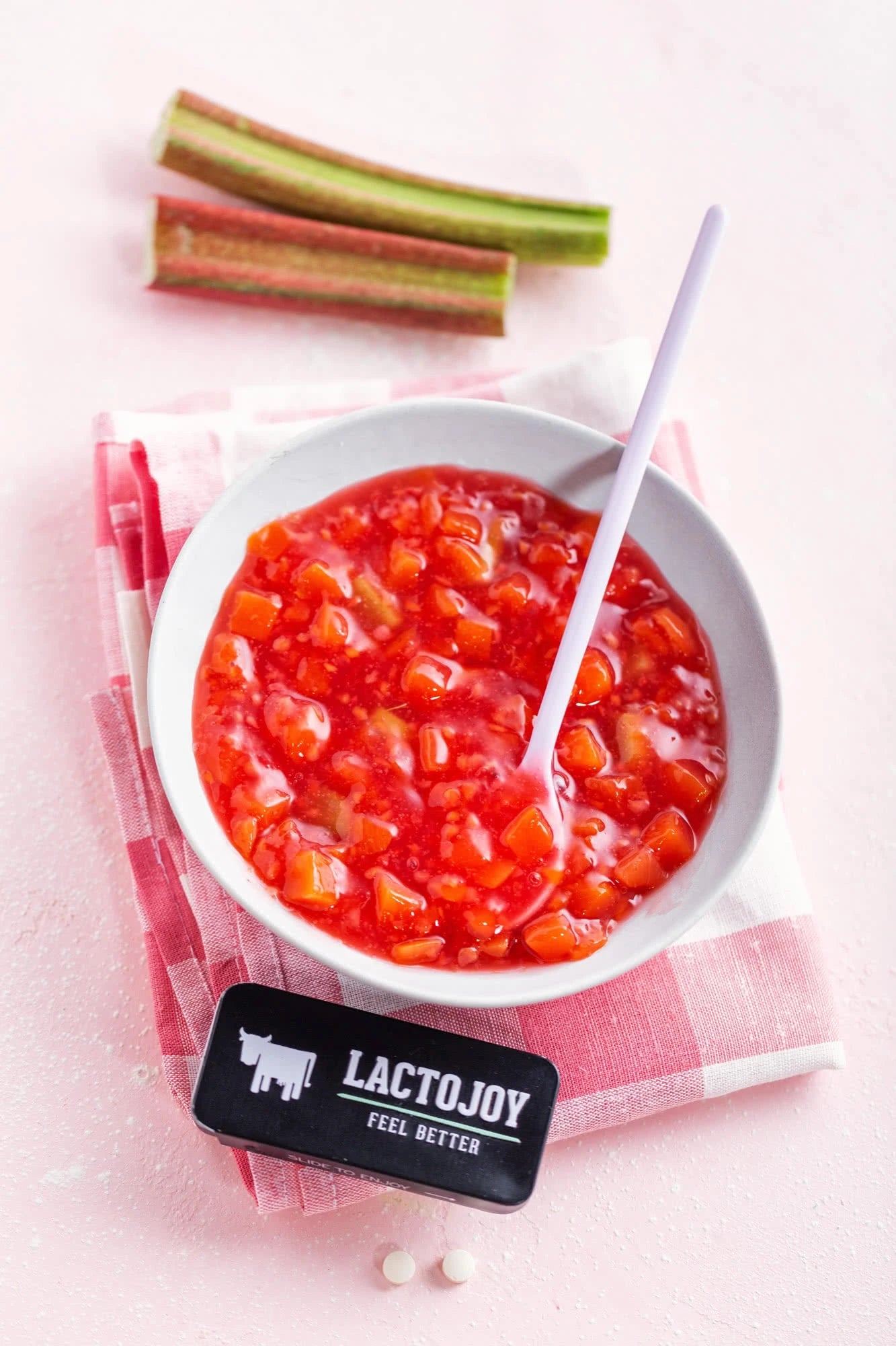 White bowl with red fruit compote, rhubarb stalks, and a LactoJoy product on a pink background